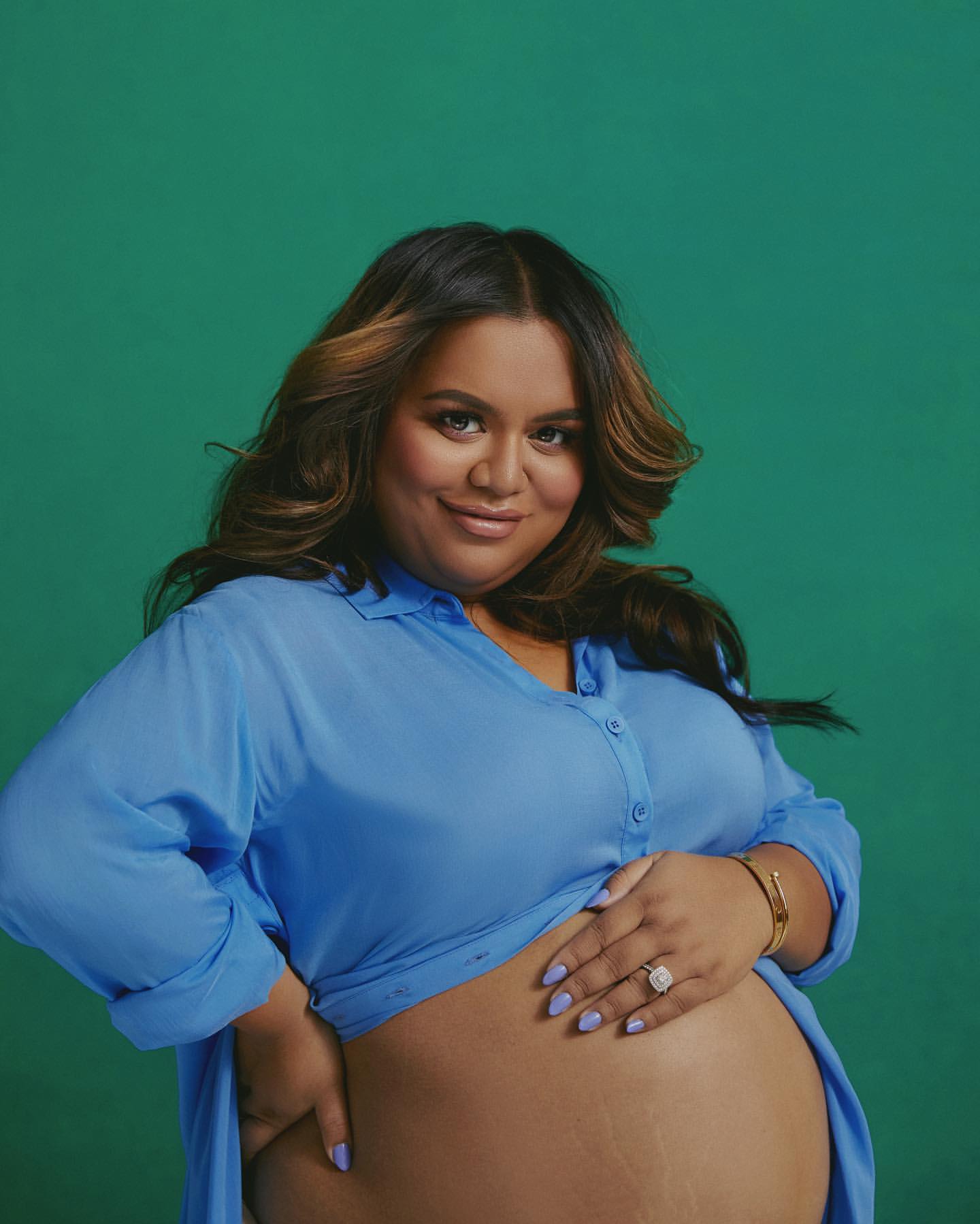 Brunette pregnant woman in a periwinkle shirt poses in front of a green backdrop, hand on belly, ring visible, smiling for a portrait.