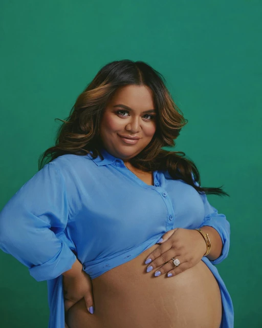 Brunette pregnant woman in a periwinkle shirt poses in front of a green backdrop, hand on belly, ring visible, smiling for a portrait.