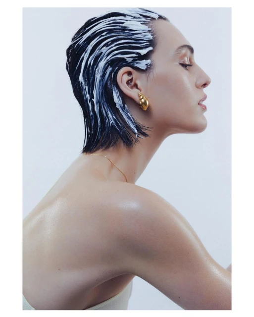 Profile of a woman with cream in her dark hair, golden earring, and bare shoulder against a pale backdrop in studio lighting.