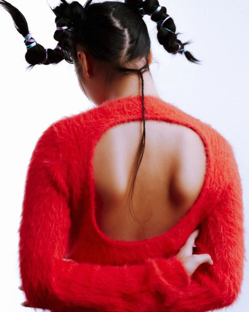 Back view of a woman with braided pigtails wearing an orange fuzzy sweater with a circular cutout exposing the back. Light background.