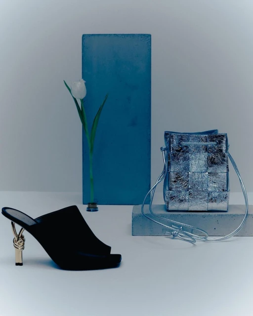 Still life featuring a black high-heeled mule shoe, a silver woven handbag, and a single white tulip against a minimalist backdrop.