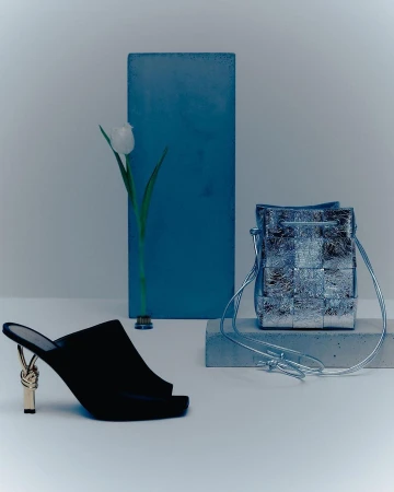 Still life featuring a black high-heeled mule shoe, a silver woven handbag, and a single white tulip against a minimalist backdrop.