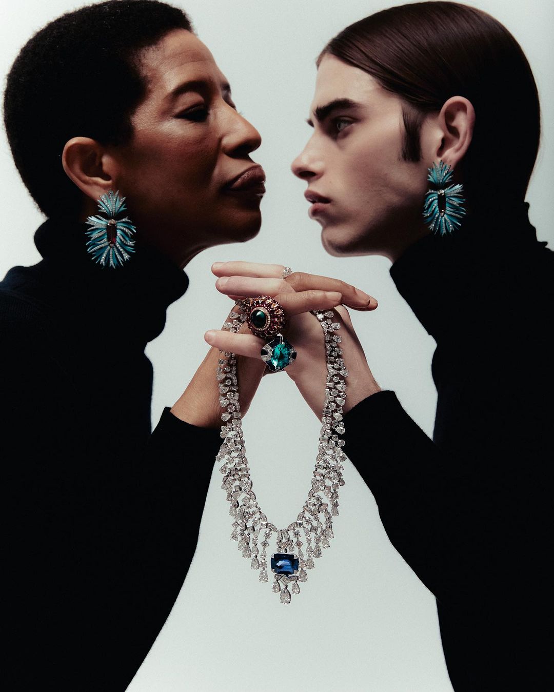 Two people in black turtlenecks display jewellery against a white background. A woman and a younger person hold a diamond necklace.