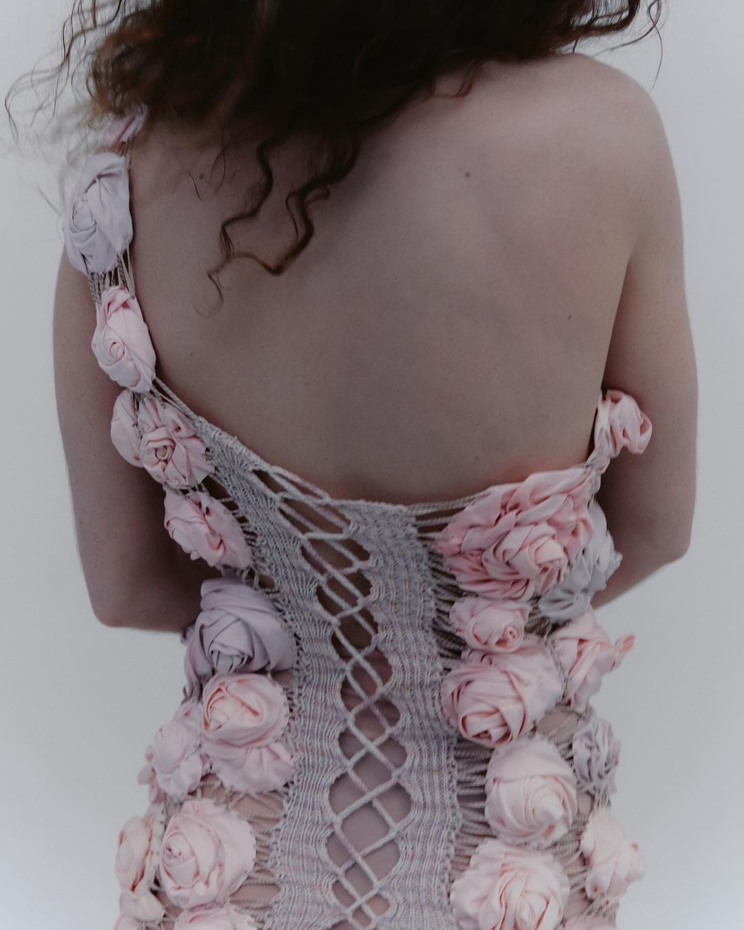 Back view of a fair-skinned adult female with curly brown hair wearing a light grey, woven dress adorned with pink fabric roses.