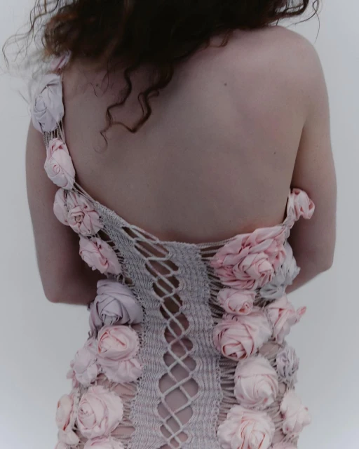 Back view of a fair-skinned adult female with curly brown hair wearing a light grey, woven dress adorned with pink fabric roses.