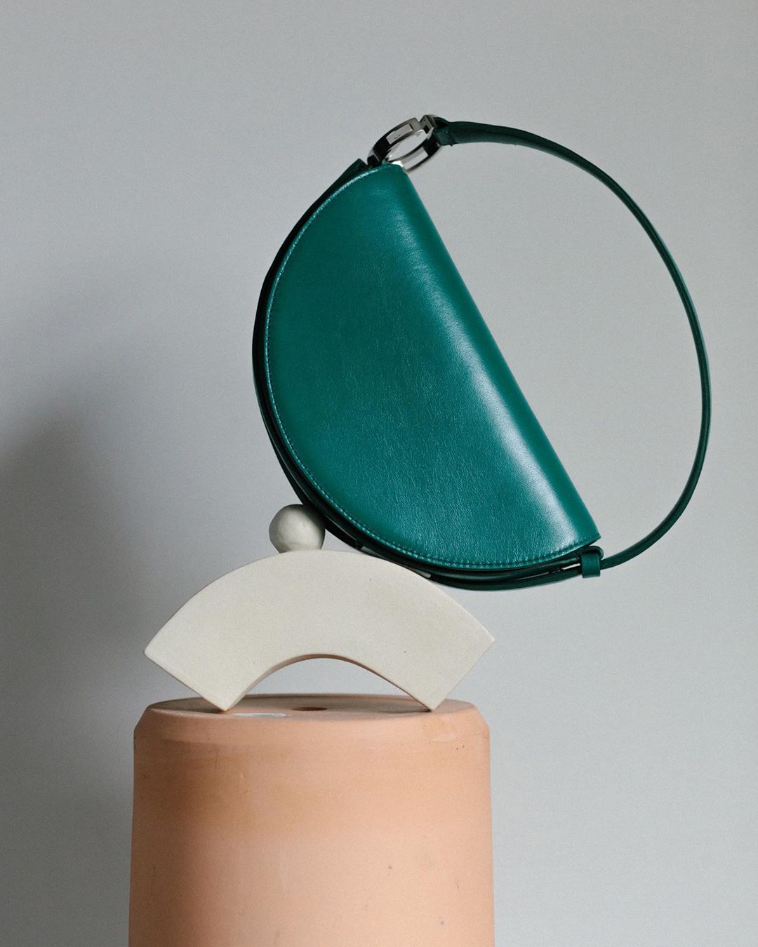 Green leather half-moon handbag is displayed on a platform with geometric shapes in a studio setting, providing a sleek product shot.