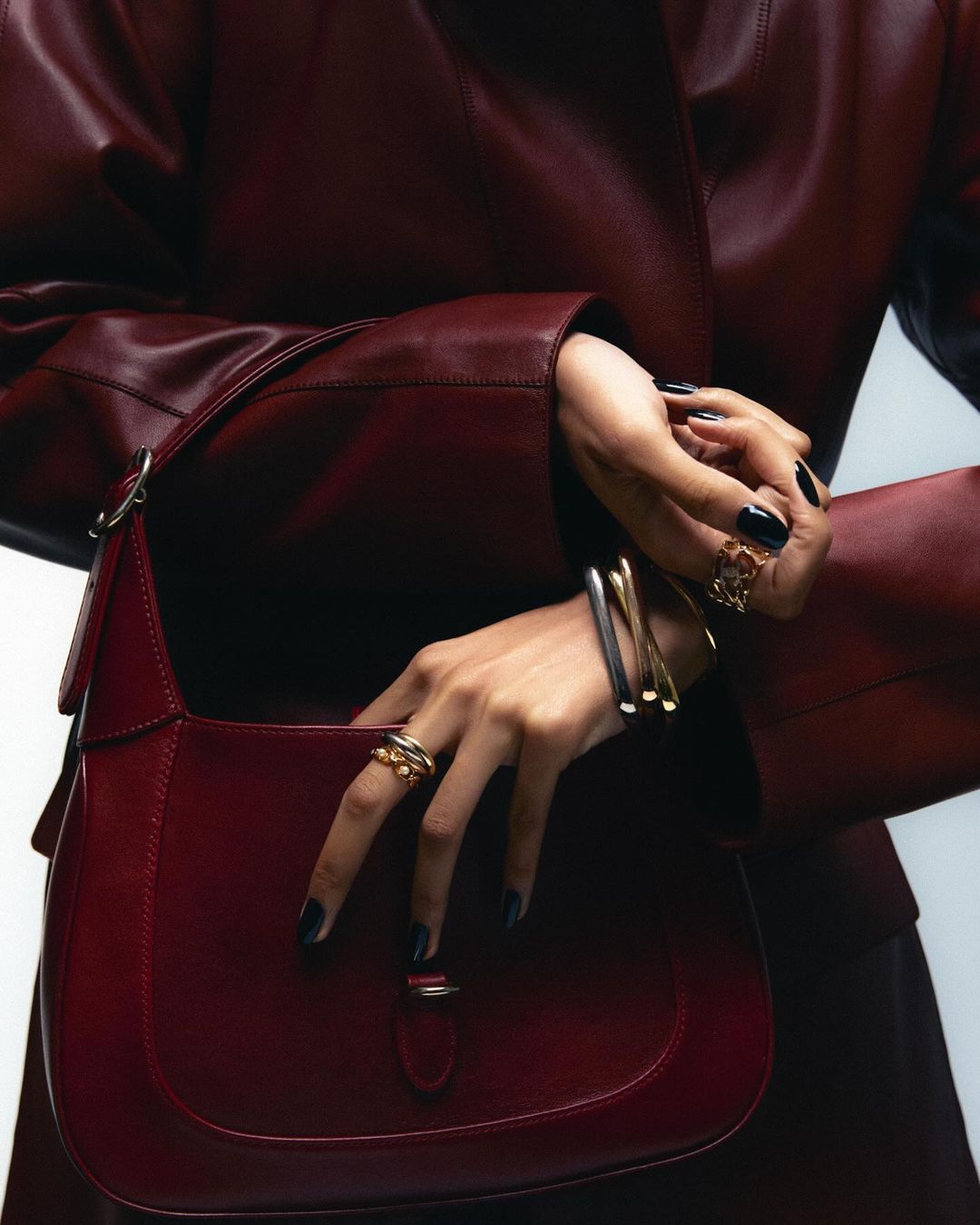 Close-up studio shot of a woman's hands with rings and bracelets holding a red leather shoulder bag, showcasing fashion accessories.