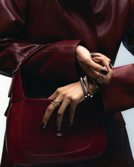 Close-up studio shot of a woman's hands with rings and bracelets holding a red leather shoulder bag, showcasing fashion accessories.
