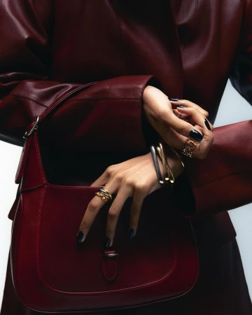 Close-up studio shot of a woman's hands with rings and bracelets holding a red leather shoulder bag, showcasing fashion accessories.