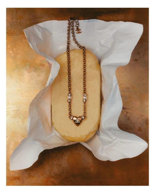 Elegant gold necklace with pearl and jewel accents draped on soft yellow cheese inside crinkled white paper on bronze background.