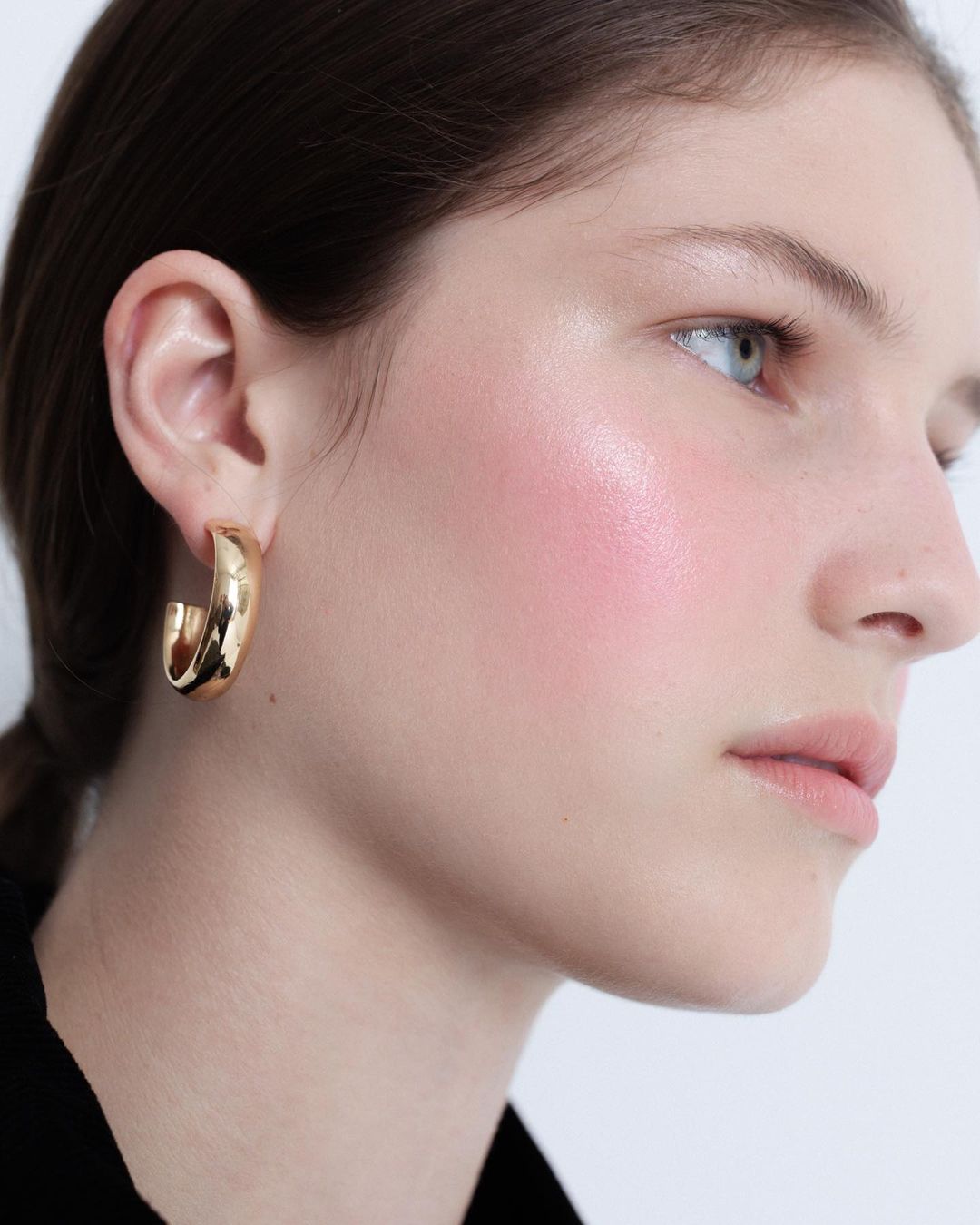 Close-up portrait of a woman wearing a thick gold hoop earring and a black top against a plain background by Vanessa Granda.