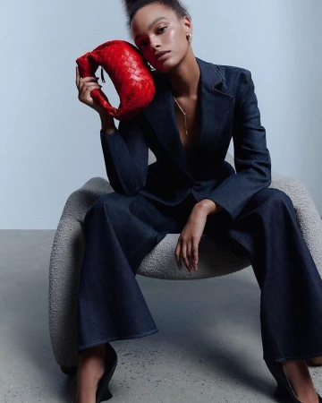 Woman in dark denim suit seated in a textured armchair holding a bright red woven handbag photographed by Vanessa Granda