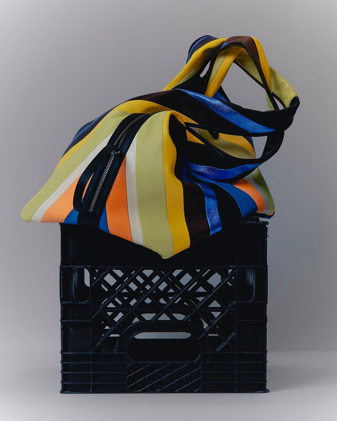 Striped tote bag with unique knotted handles sits atop a black milk crate against a light gray backdrop in a studio shot.