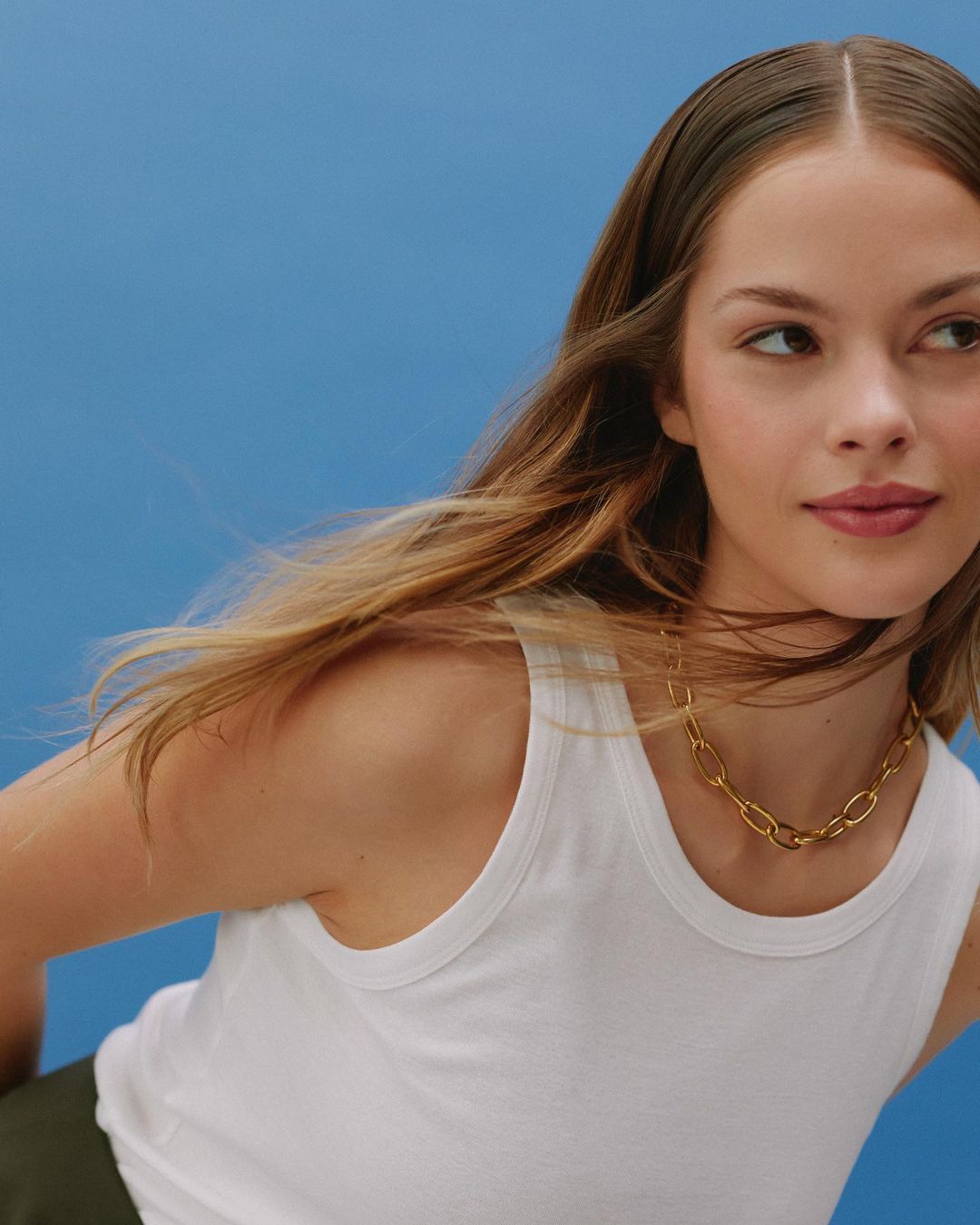 Portrait of a woman wearing a white tank top and a gold chain necklace against a solid blue background