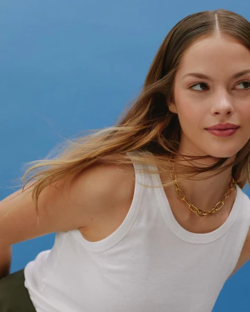 Portrait of a woman wearing a white tank top and a gold chain necklace against a solid blue background