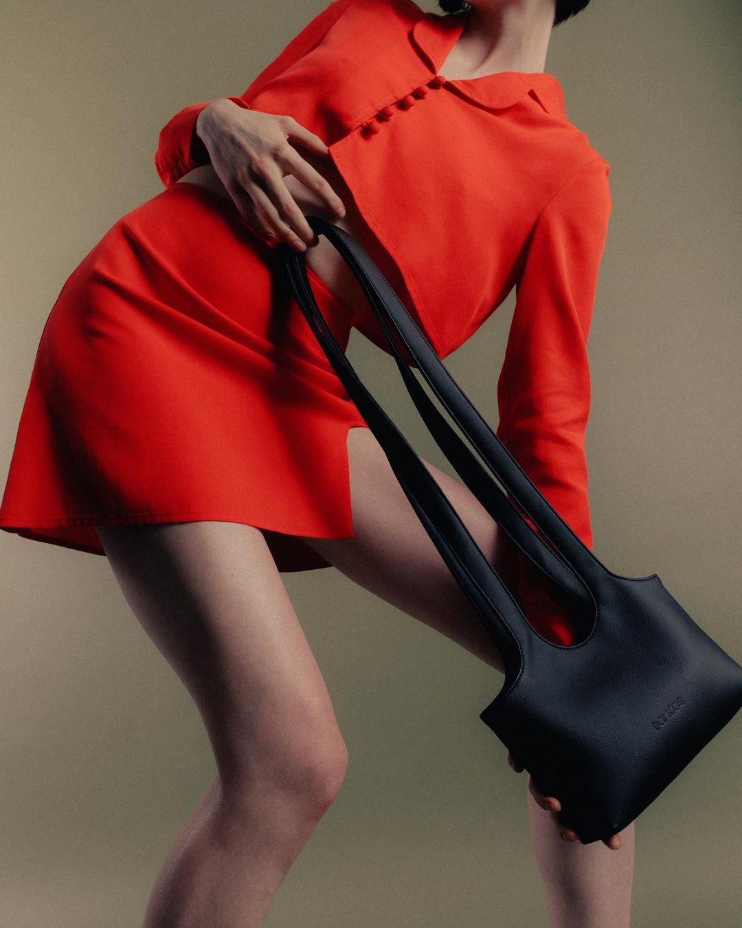 Woman in red dress holds black leather handbag in studio setting, emphasizing fashion and accessory design with a focus on the bag.