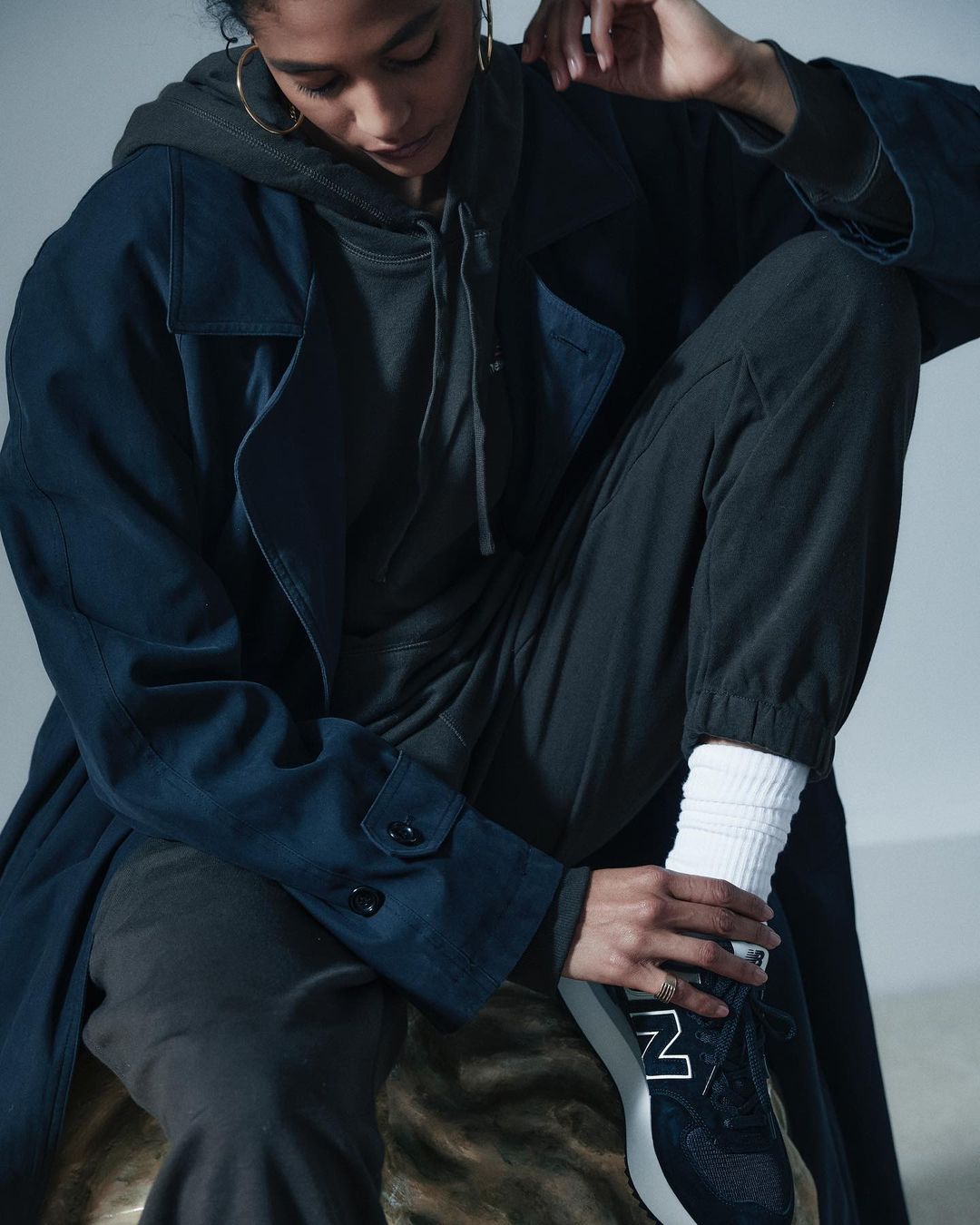 Woman seated in a navy trench coat and gray hoodie, ties her black New Balance sneaker in a studio setting with soft, diffused lighting.