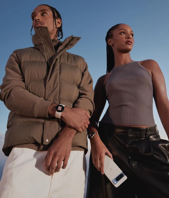 Two models pose outdoors with a phone and smartwatch against a blue sky, showcasing modern tech and minimalist fashion.