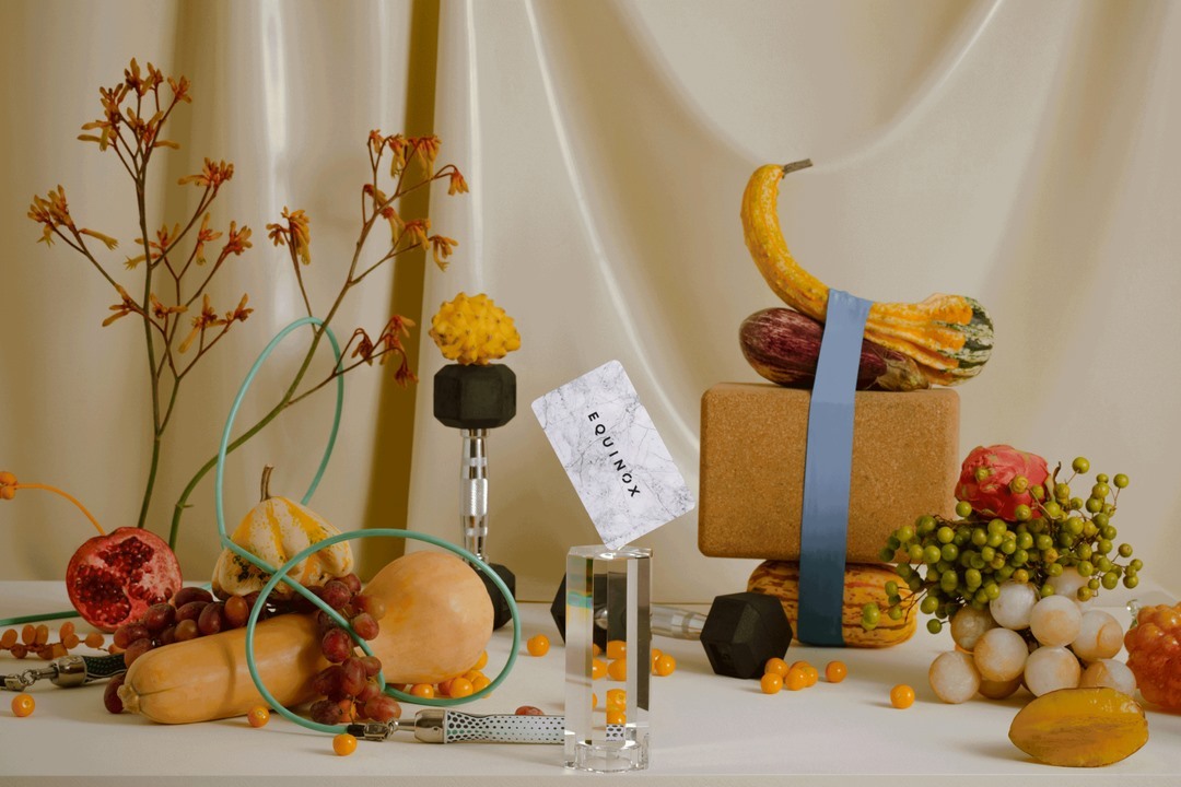Still life of fitness equipment and fruits with autumnal colors, featuring dumbbells, gourds, grapes, jump rope, and an Equinox card.