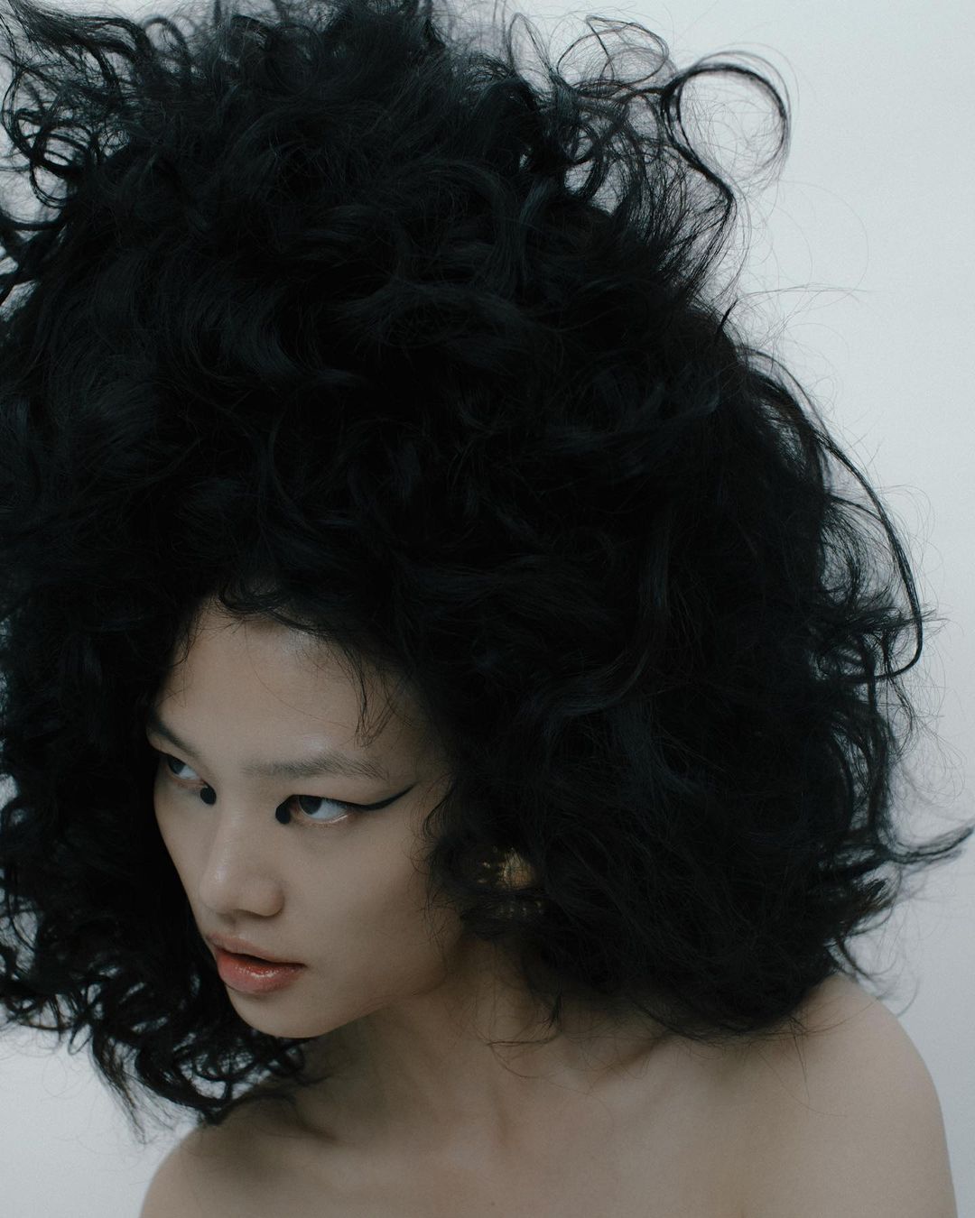 A portrait photograph of an Asian woman with large, curly black hair, dramatic black eyeliner, bare shoulders, and neutral background.