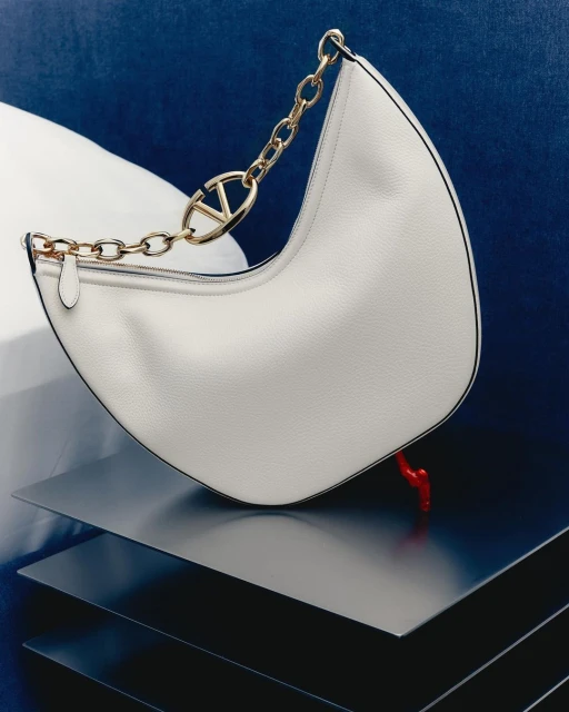 Studio shot of a white leather hobo bag with a gold chain handle and logo, placed on geometric metallic platforms against a blue background.