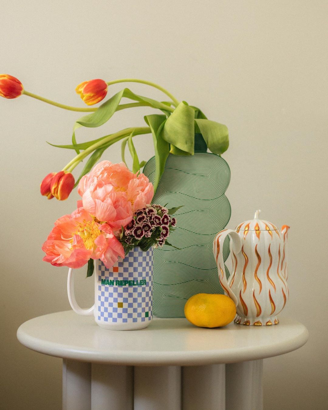Still life composition featuring vibrant tulips and peonies in a checkered mug, alongside a decorative vase, teapot, and orange on a white table.