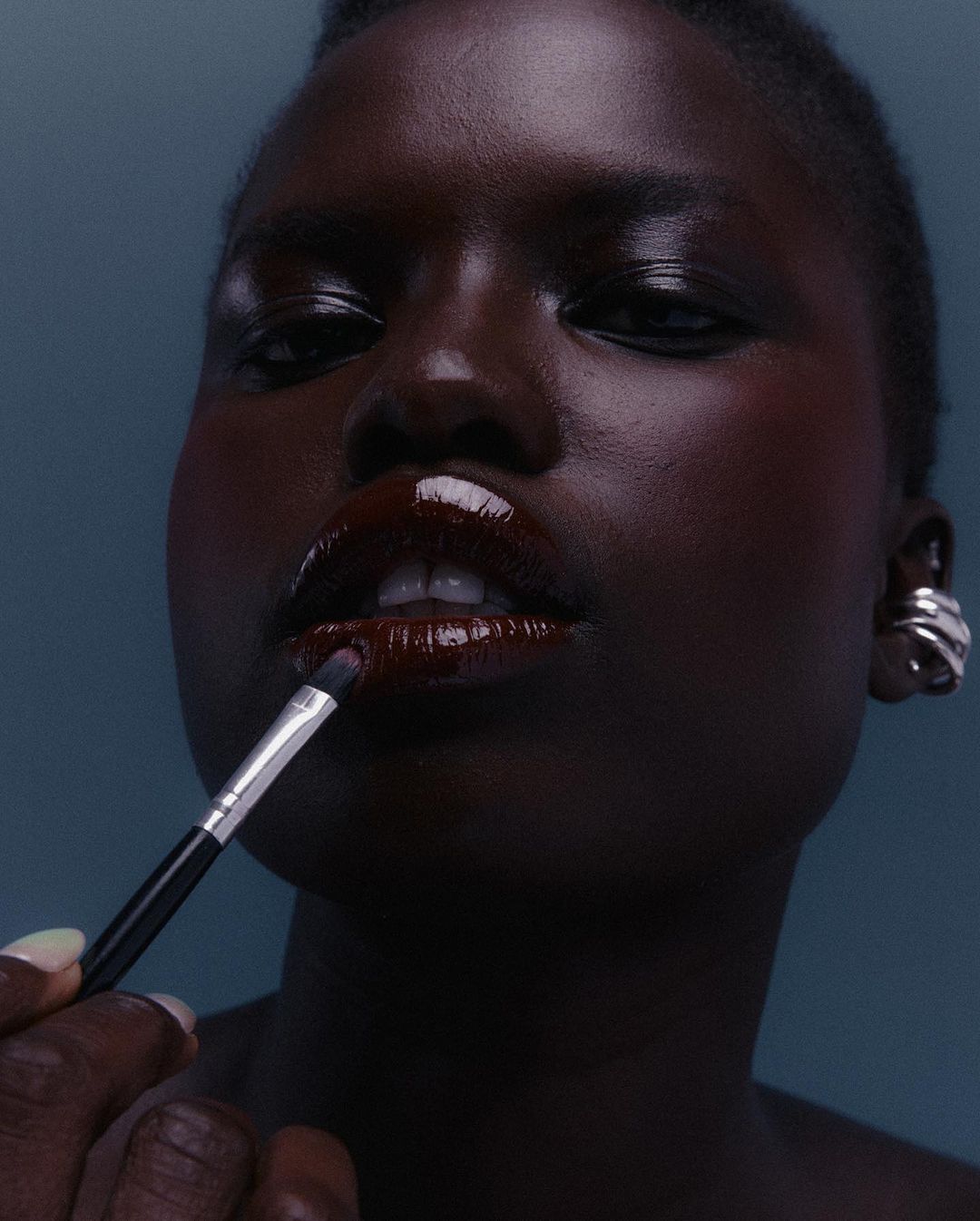 Close-up of a Black woman applying dark lip gloss with a makeup brush. Focus on her lips and the makeup brush, against a blue background.
