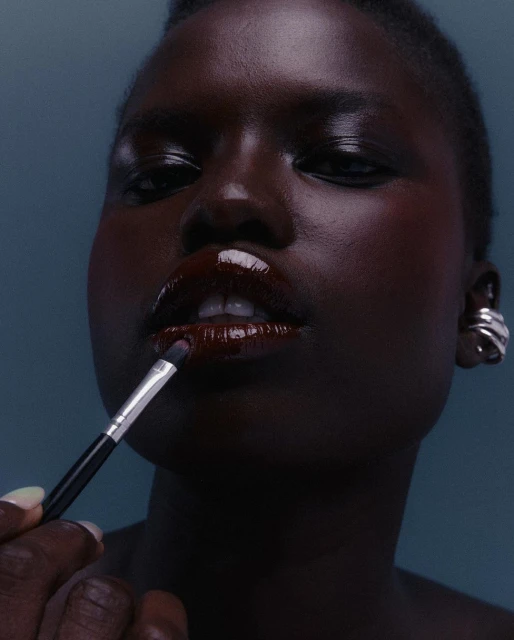 Close-up of a Black woman applying dark lip gloss with a makeup brush. Focus on her lips and the makeup brush, against a blue background.
