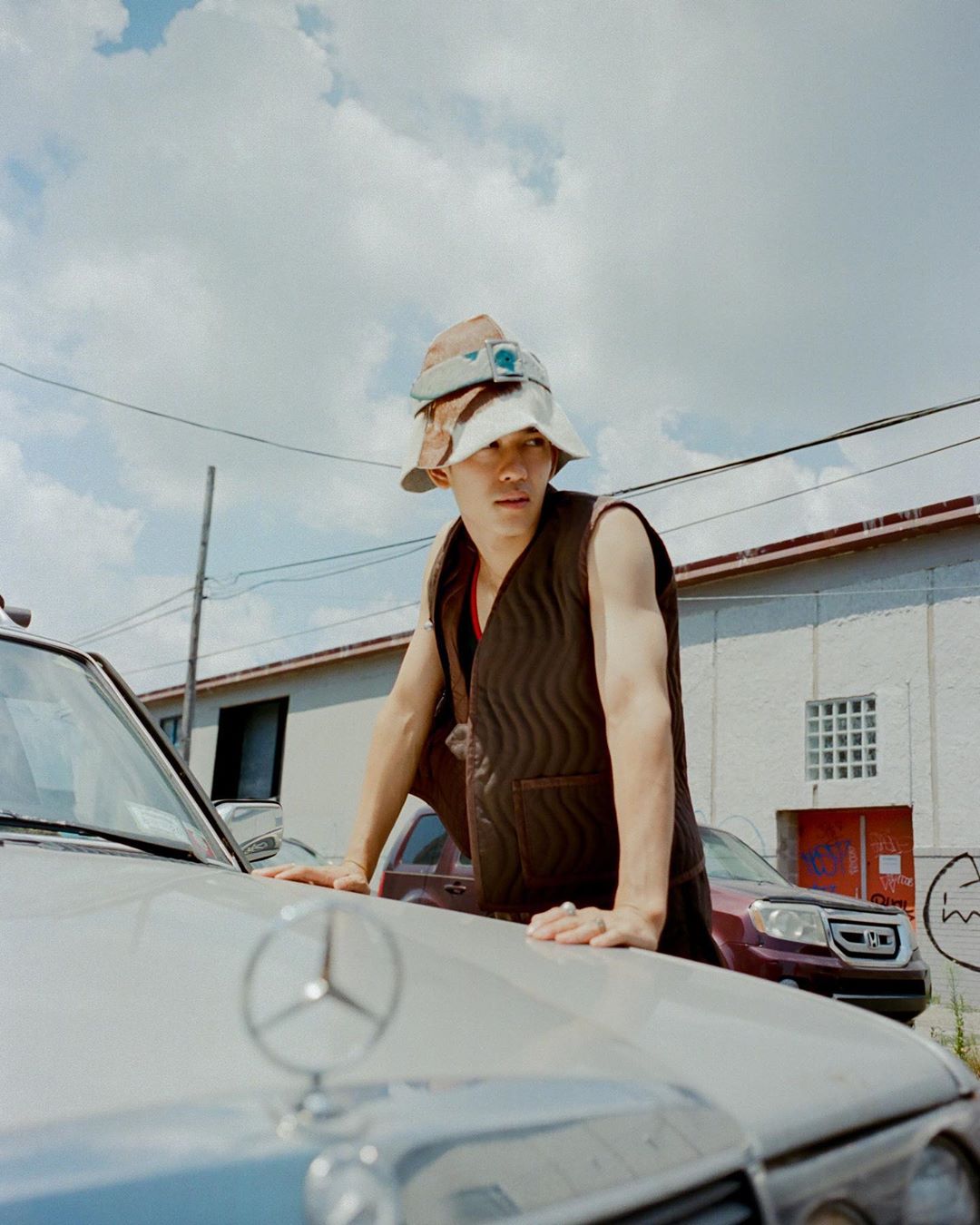 Young man in bucket hats and vest leans on a silver Mercedes-Benz under a cloudy sky with industrial building. Street style fashion.