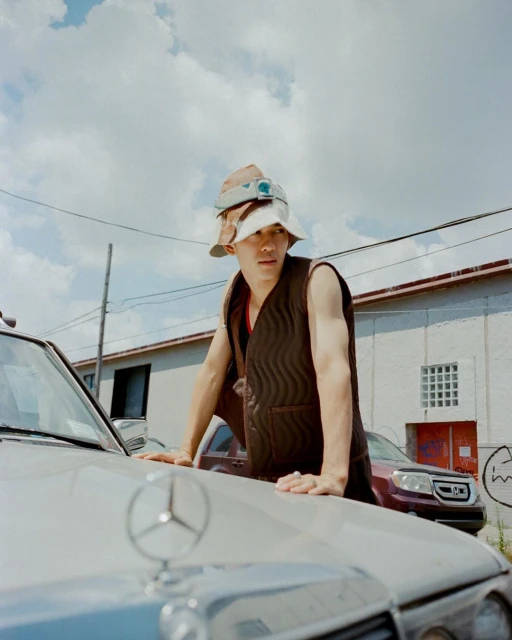 Young man in bucket hats and vest leans on a silver Mercedes-Benz under a cloudy sky with industrial building. Street style fashion.
