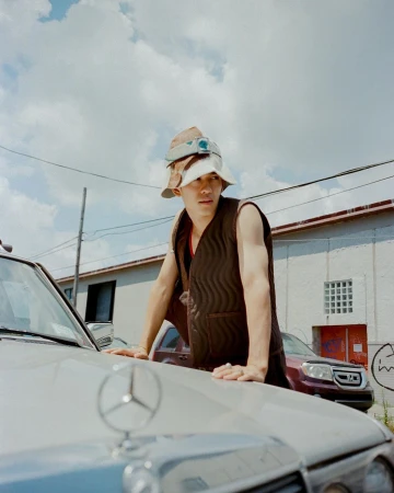 Young man in bucket hats and vest leans on a silver Mercedes-Benz under a cloudy sky with industrial building. Street style fashion.