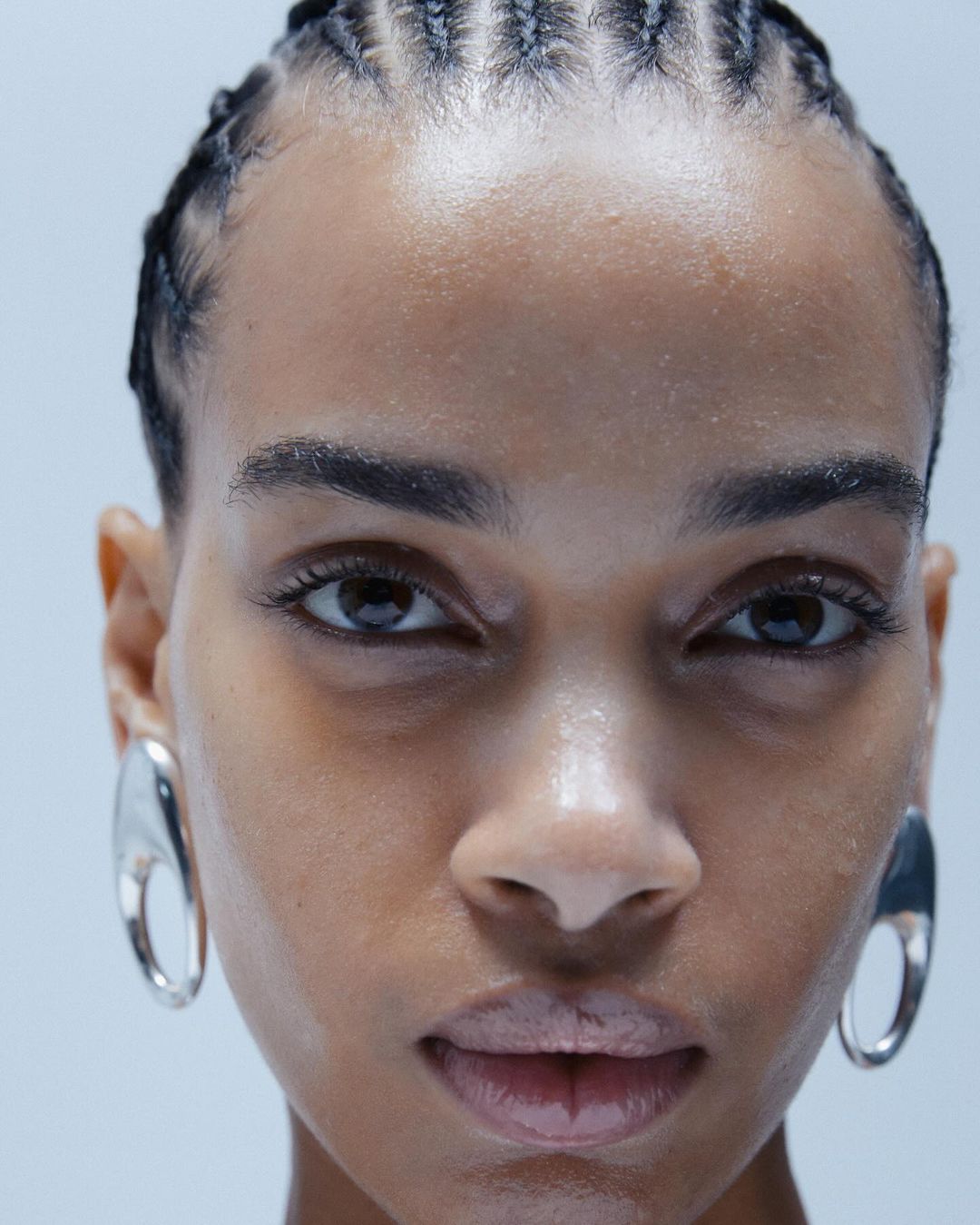 Extreme close-up portrait of a woman with braided hair wearing large silver sculptural earrings against a plain light background