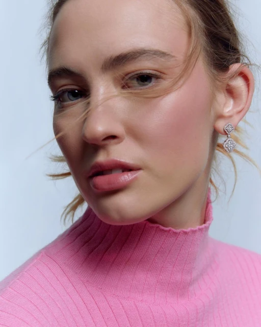 Close-up of a fair-skinned young woman with blue eyes wearing a pink ribbed turtleneck and diamond flower earrings against a white backdrop.
