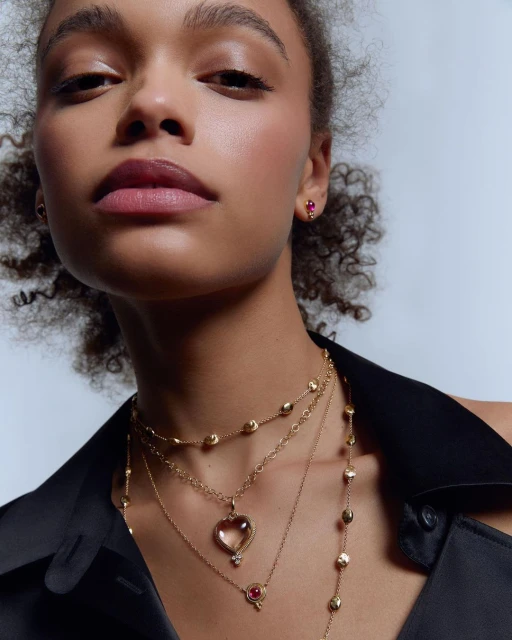 Adult woman with curly hair wearing multiple gold necklaces and a gold stud earring in a studio setting, fashion photography.