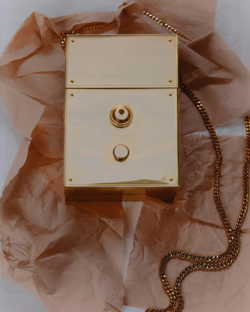 Still life shot of a gold metallic box purse with a chain strap, laying on a tan crumpled paper surface in a studio setting.