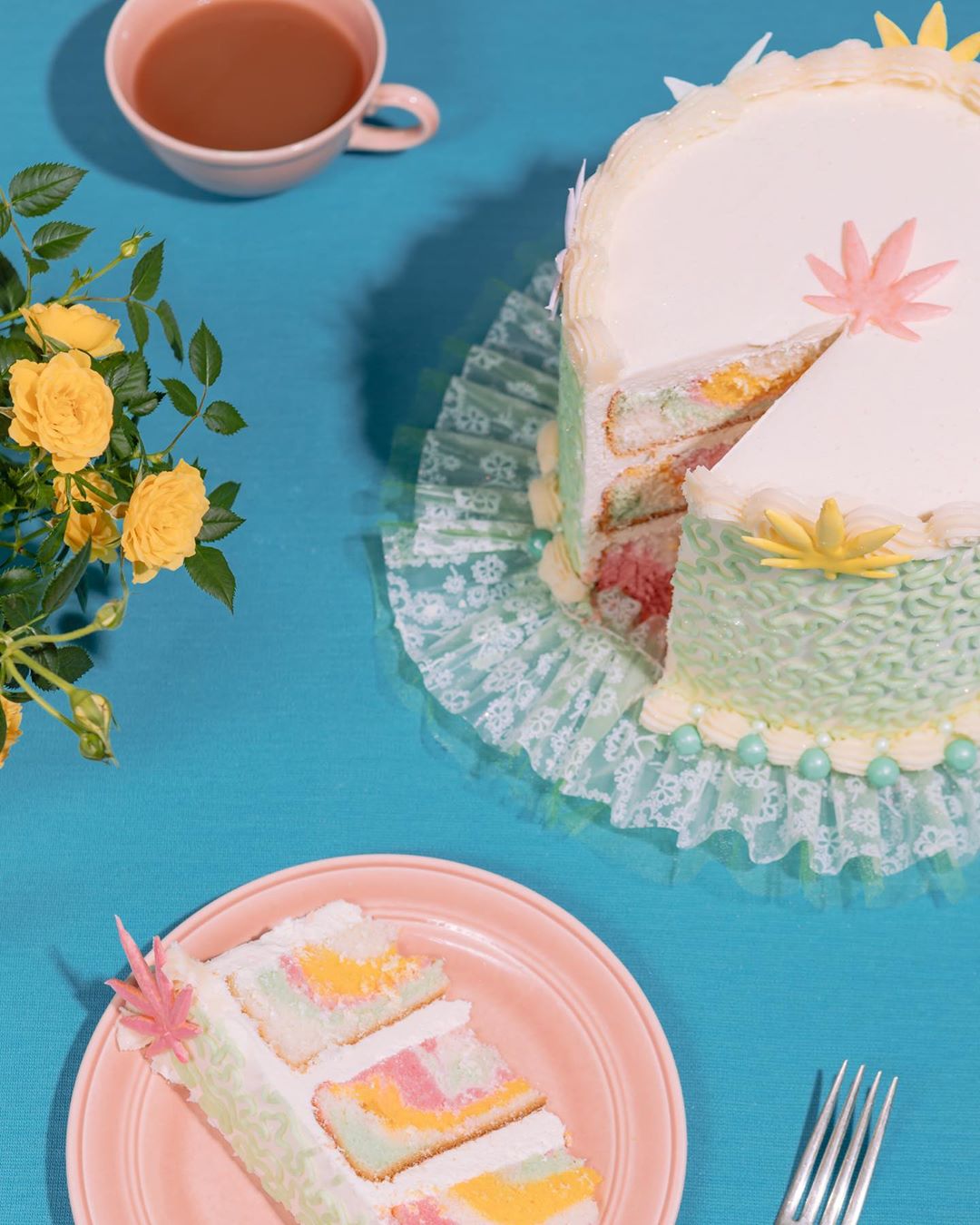 Overhead shot of a sliced pastel layer cake on a pink plate, with a fork, flowers, a cup of tea, all on a teal background.