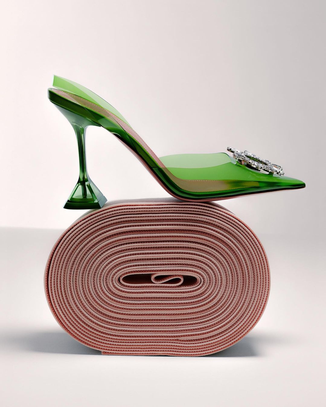 Elevated view of a stylish transparent green high-heel shoe with rhinestone embellishment on a spool of pink elastic.