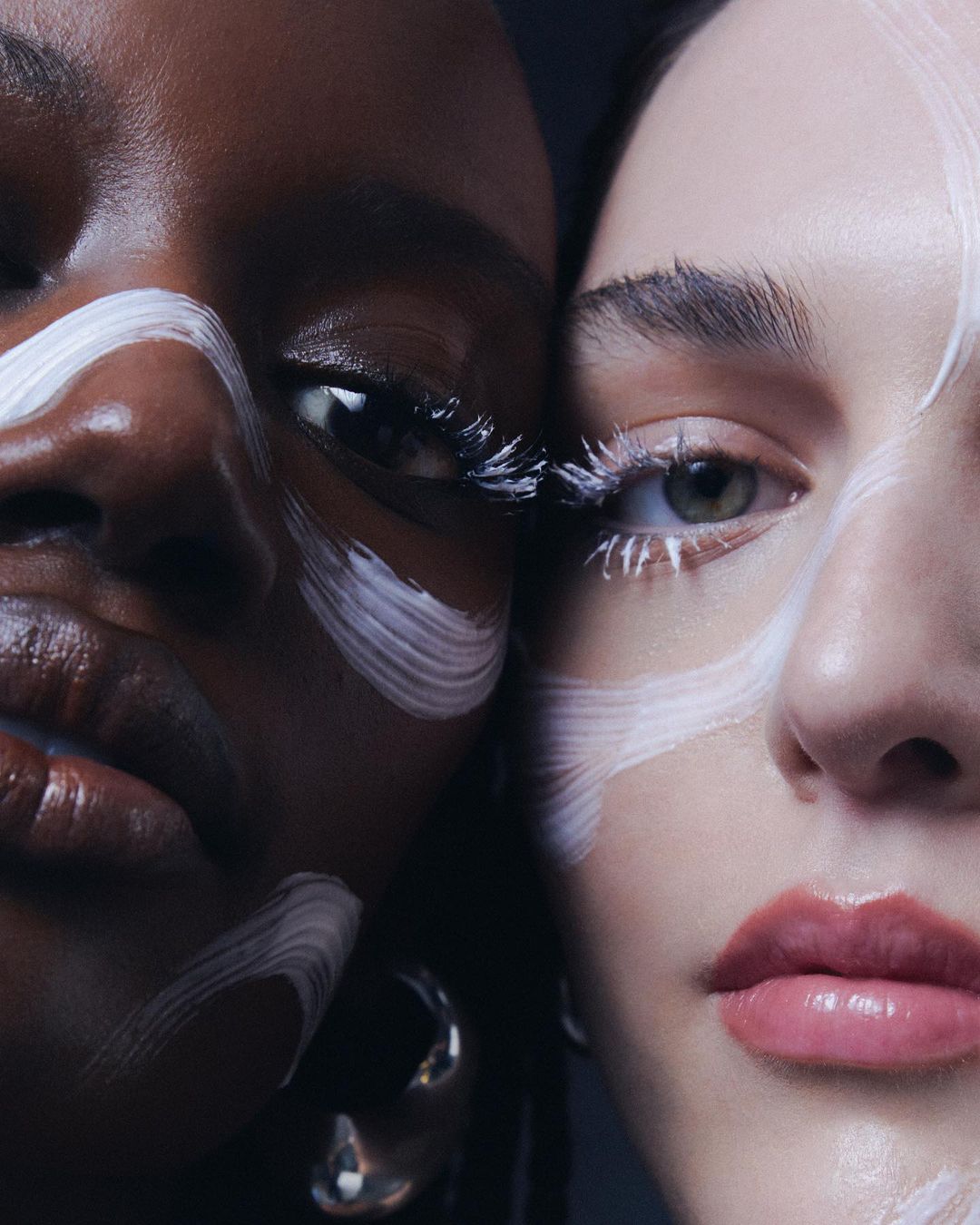 Close-up studio shot of two adult female faces with makeup and matching silver hoop earrings; stark white paint across their faces.
