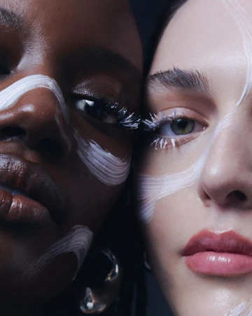 Close-up studio shot of two adult female faces with makeup and matching silver hoop earrings; stark white paint across their faces.