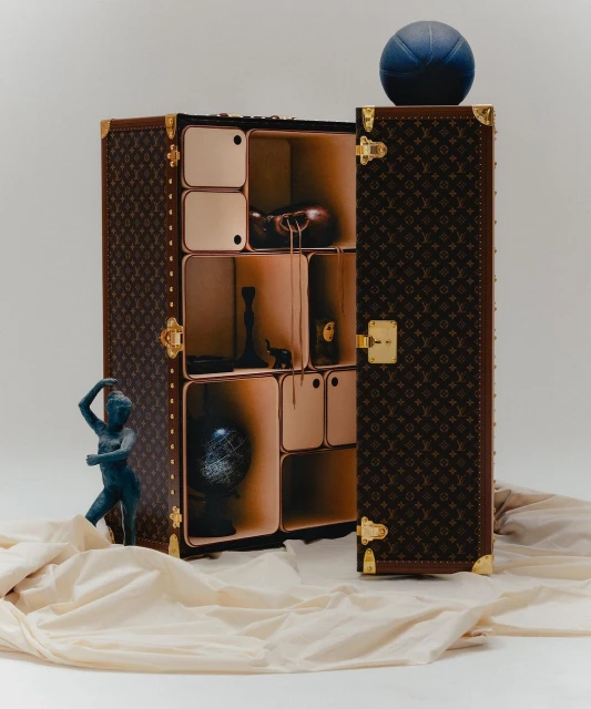 Luxury Louis Vuitton trunk with open door reveals shelves displaying decorative objects, a basketball sits on top, draped cloth at base.
