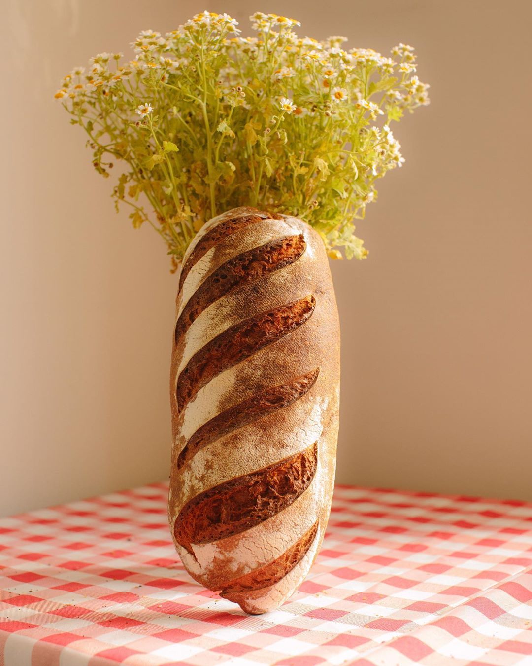Rustic sourdough bread loaf with striped pattern as a vase, holding a bundle of chamomile flowers on a red checkered tablecloth.