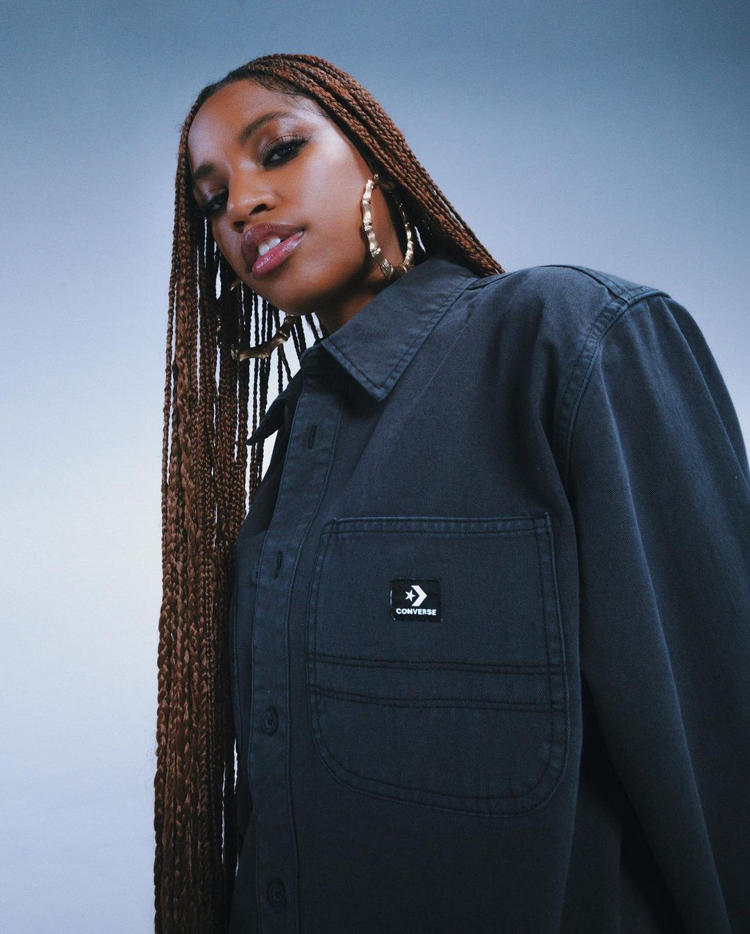 Close-up of a woman wearing a black jacket with gold hoop earrings, looking directly at the camera. She has long braided hair.