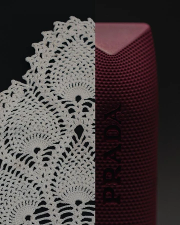 Close-up studio shot features a white crocheted doily and a maroon-colored PRADA speaker against a dark background.