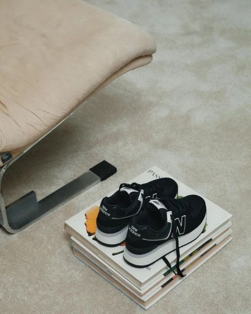 Black New Balance sneakers sit atop stacked books next to a modern chair, creating a minimalist and stylish indoor setting.