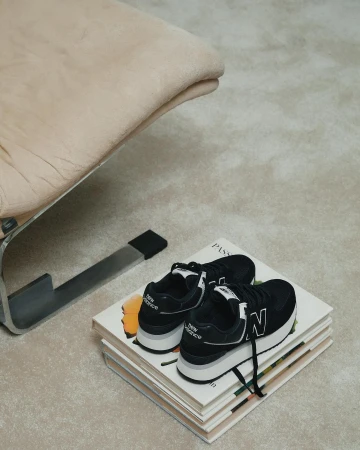 Black New Balance sneakers sit atop stacked books next to a modern chair, creating a minimalist and stylish indoor setting.