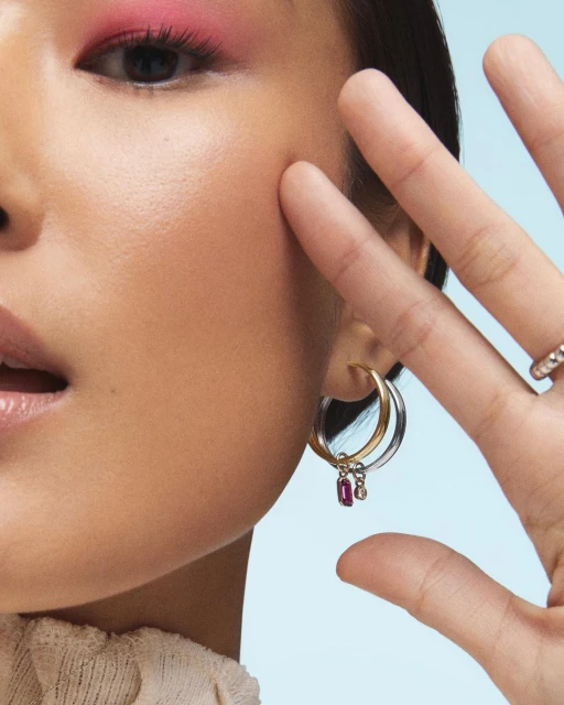 Close-up of a woman showcasing hoop earrings, with pink eyeshadow and a neutral top against a soft blue backdrop.