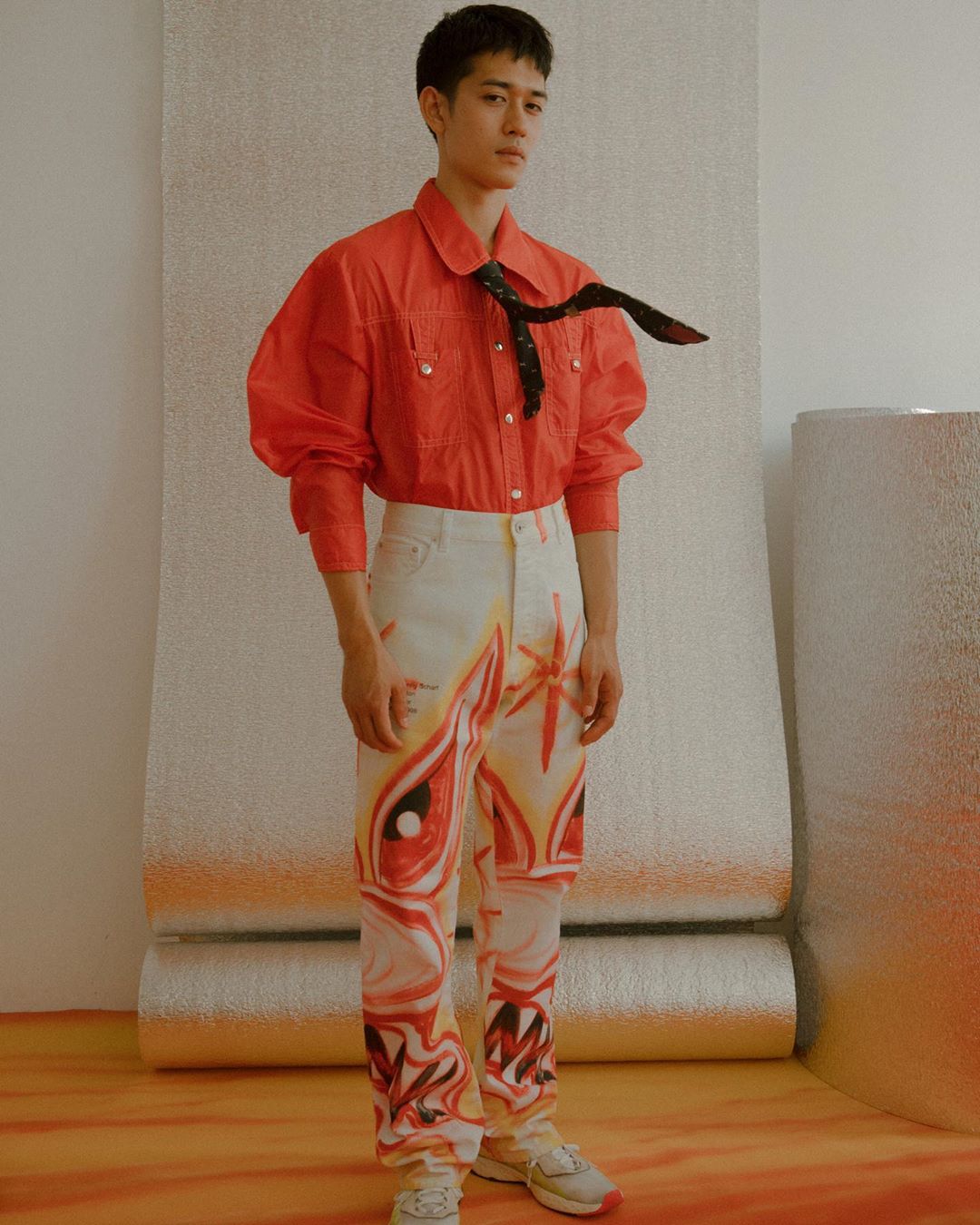 Young Asian man poses in a red shirt and artistically painted jeans against a silver backdrop. Fashion editorial shot indoors.