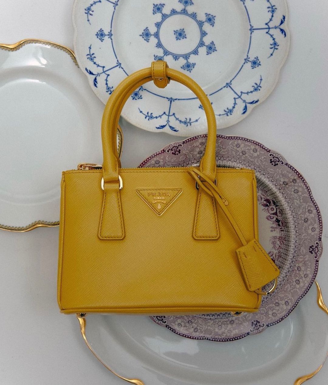 A mustard yellow Prada handbag is centered against a backdrop of three decorative plates with scalloped rims and blue designs.