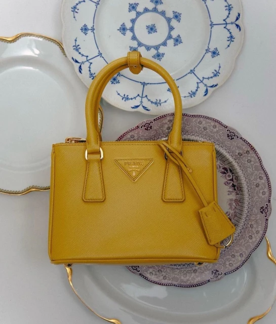 A mustard yellow Prada handbag is centered against a backdrop of three decorative plates with scalloped rims and blue designs.