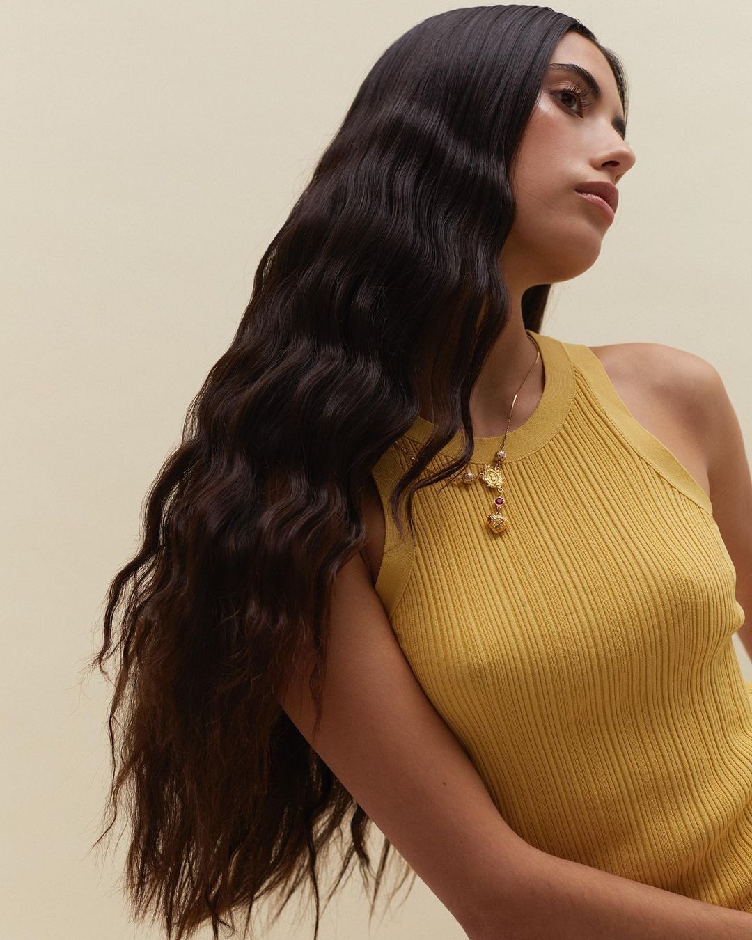 Portrait of a young adult woman with long dark hair, wearing a mustard yellow top and a gold necklace, against a neutral background.